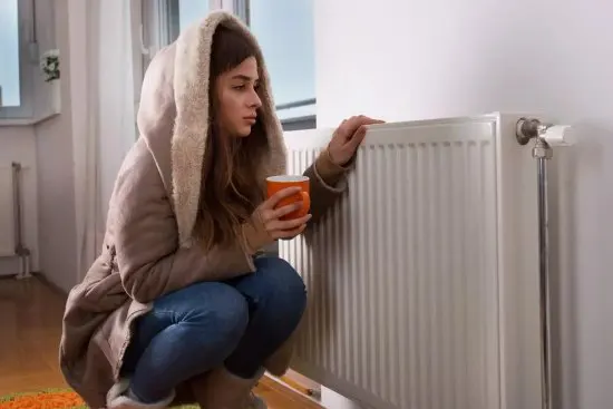 Meisje in jas leunt tegen een radiator vanwege slecht functionerende ketel door kalkaanslag.